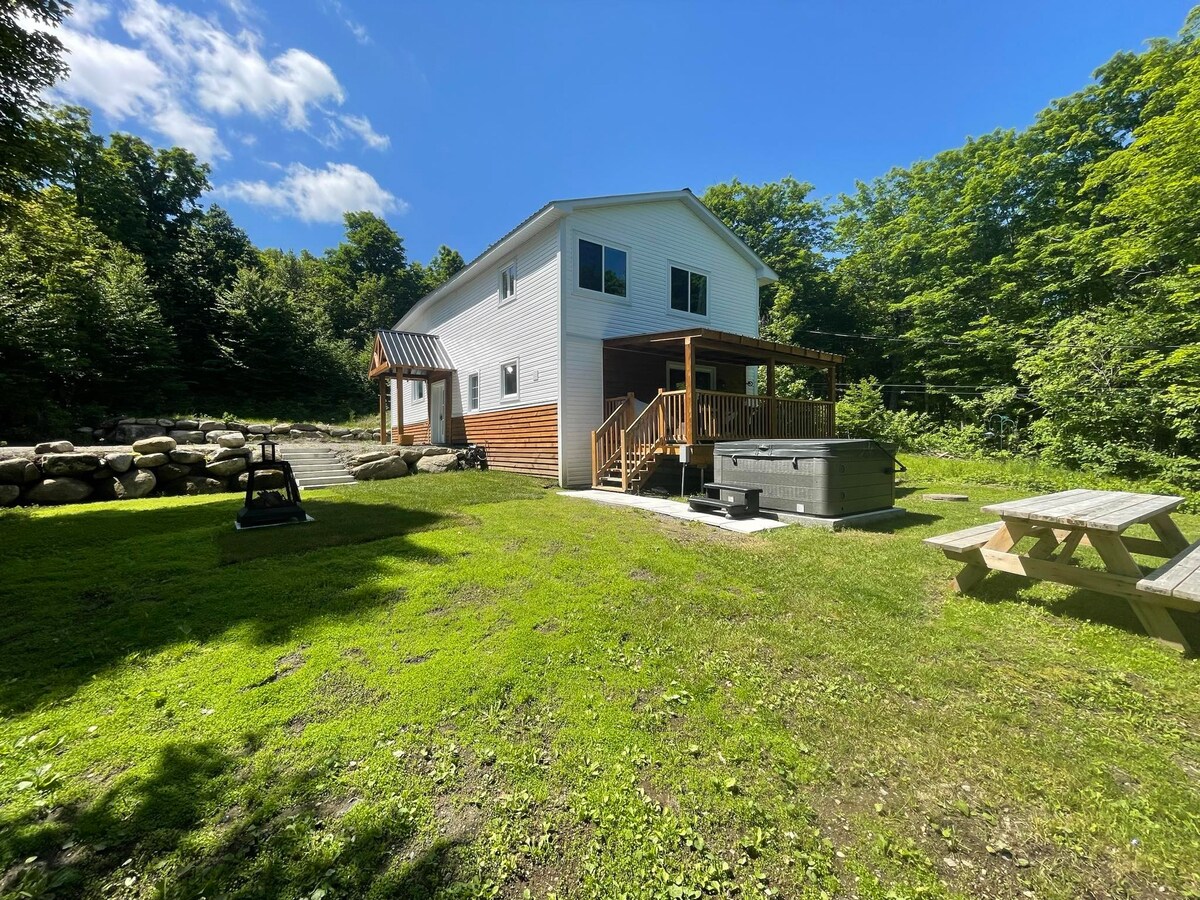 A spacious chalet is set against a backdrop of greenery, featuring a large deck with steps leading down to an open grassy area. A picnic table and a hot tub are visible in the outdoor space, complemented by stone landscaping near the structure.