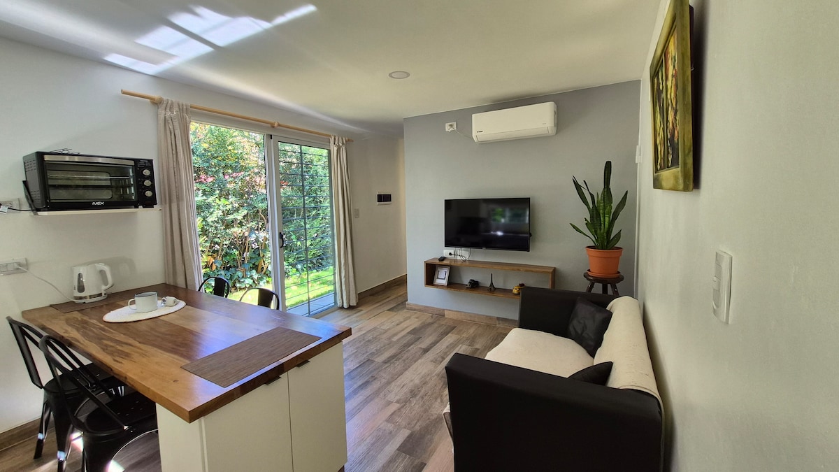 An inviting living space is featured, showcasing a modern design with a comfortable sofa and a mounted TV. A dining area with a wooden counter and chairs provides a functional gathering spot. Large glass doors lead to a lush green outdoor area, enhancing the connection with nature.