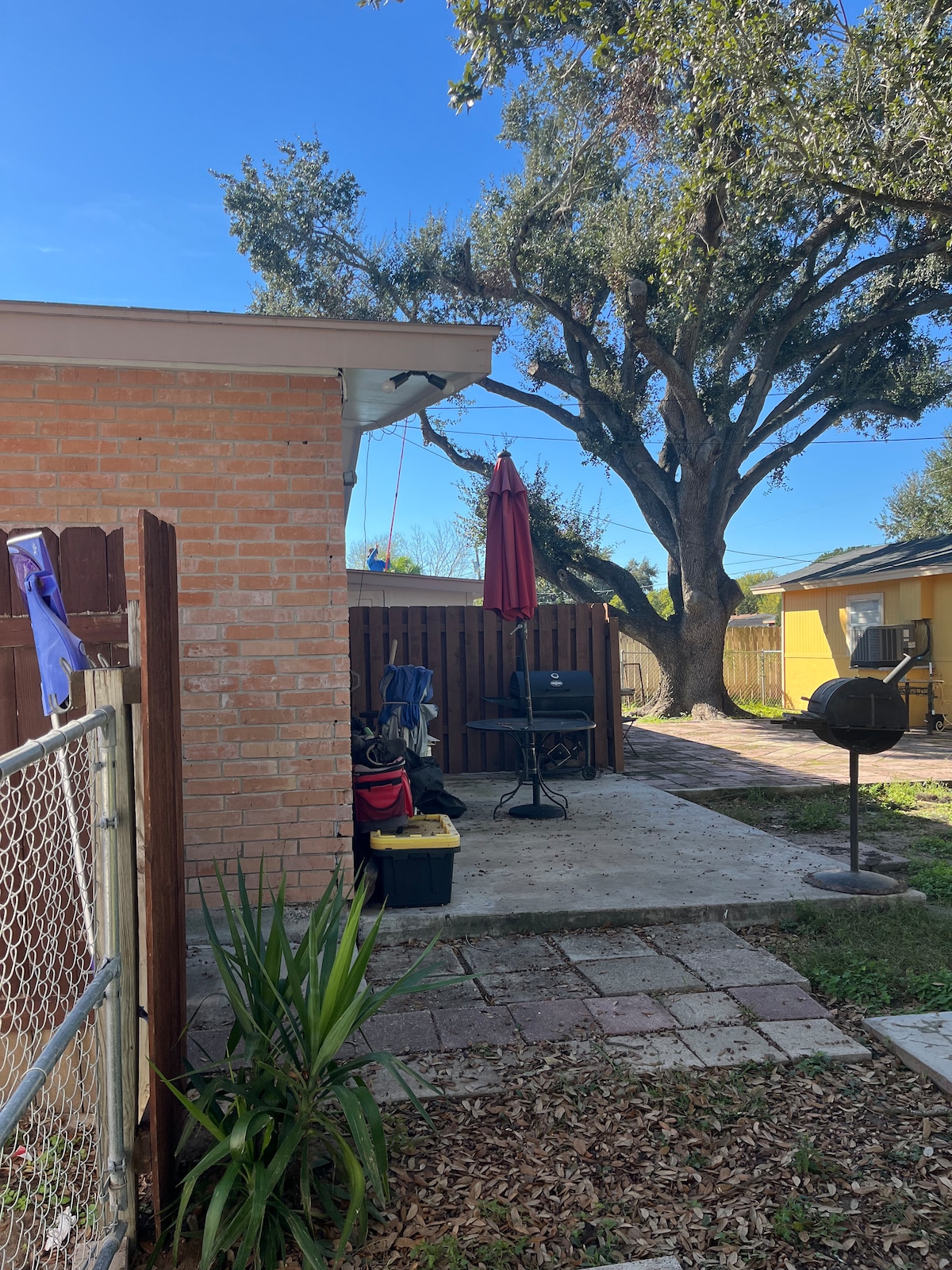 An outdoor space is depicted featuring a patio area with a round table and an umbrella. A grill is positioned beside the patio on a paved surface. Lush greenery, including an ornamental plant, is present along the edges, with a large tree providing shade in the background.