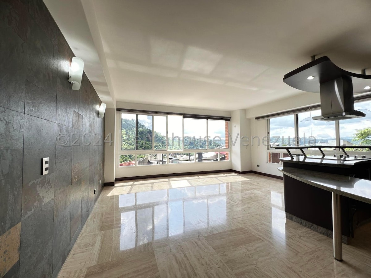 An open-concept living area is highlighted by large windows that allow natural light to fill the space. A modern kitchen is positioned to one side, showcasing sleek design elements. The marble flooring adds a polished touch, complementing the views of the surrounding landscape.