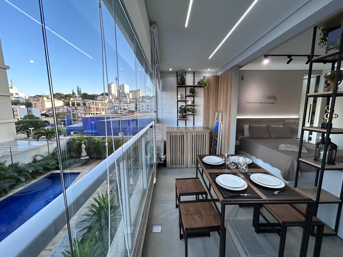 A modern dining area features a wooden table set for four, with decorative plates and glassware. A large window offers views of the surrounding city, including a glimpse of a pool and greenery. Soft lighting illuminates the space, enhancing its contemporary design.