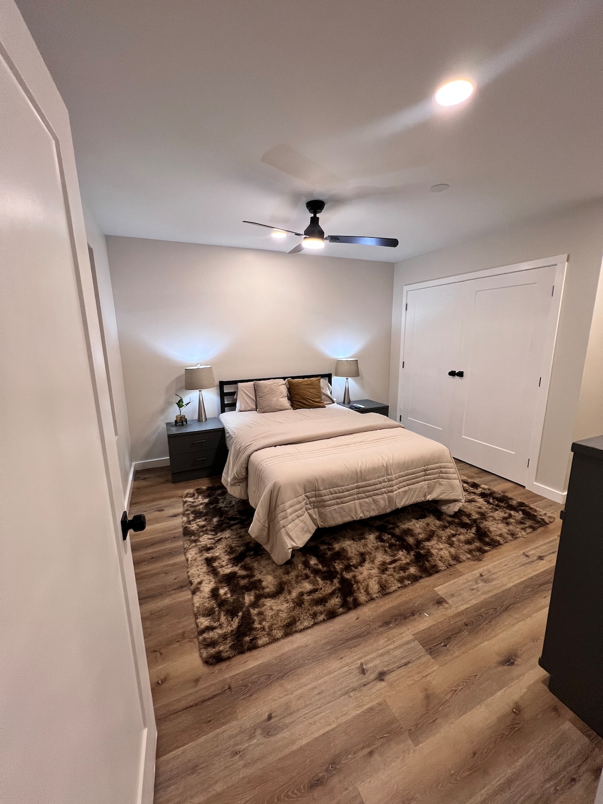 A neatly arranged bedroom features a queen bed with a light-colored blanket. Two bedside lamps illuminate the space on either side of the bed. A plush, brown area rug adds texture to the floor, while light walls and a ceiling fan contribute to the room's airy feel.