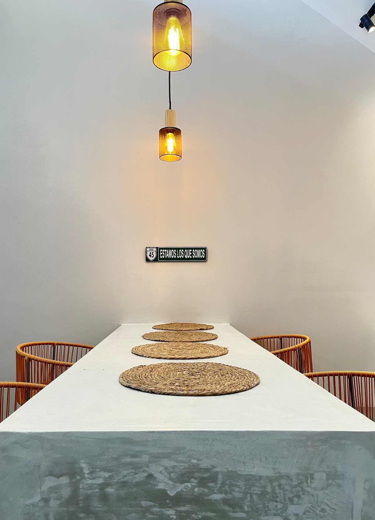 A long, minimalist dining table with textured placemats is featured, accompanied by two round rattan chairs. Two pendant lights hang above, casting a warm glow on the simple, neutral-toned walls, creating an inviting space for dining.
