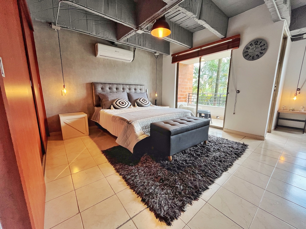 A modern bedroom is presented with a spacious bed featuring patterned pillows. A cozy area rug adds texture to the floor, while a large window provides natural light. An air conditioning unit is positioned above the bed. The room includes a closet and a stylish seating area.