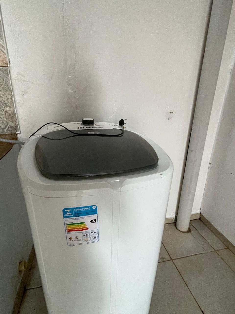 A white washing machine is positioned against a clean, tiled wall. Its grey lid is slightly raised, revealing the control panel on top. The surrounding area is well-lit, highlighting the appliance's modern design.