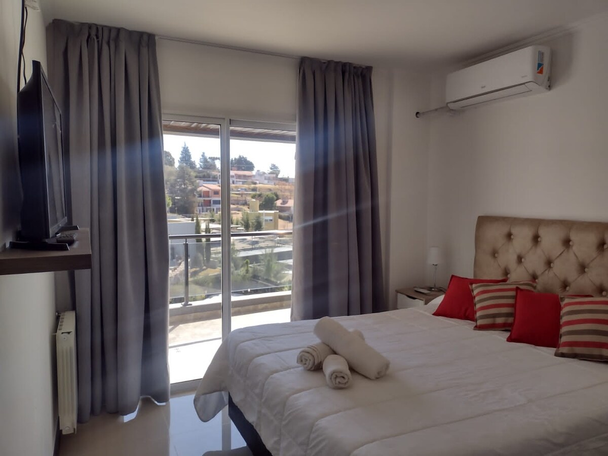A bright bedroom features a spacious, neatly made bed with decorative pillows. Natural light enters through sliding glass doors that lead to a private balcony. Soft, neutral curtains frame the window, while a mounted television and bedside lamp add functionality.