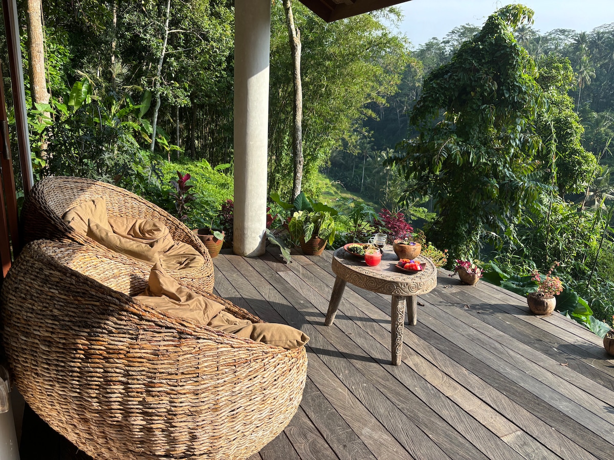A spacious deck features two large woven chairs with soft cushions, complemented by a natural wooden table. Surrounded by lush greenery, the setting offers unobstructed views of the vibrant jungle landscape, enhancing relaxation in a serene outdoor environment.