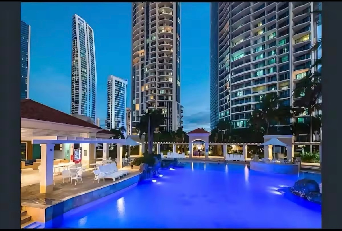 An expansive outdoor pool is illuminated in soft blue light, surrounded by high-rise buildings. Lounge chairs and covered cabanas provide seating areas, while a shaded area with a dining table and chairs is visible in the background, creating a welcoming outdoor space.