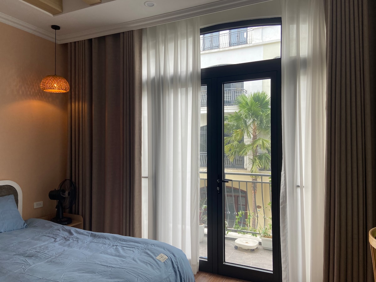 A stylish bedroom features a spacious bed with a soft blue duvet. Tall windows are framed by sheer and solid curtains, allowing natural light to fill the room. A balcony view shows green palm trees, enhancing a sense of tranquility.