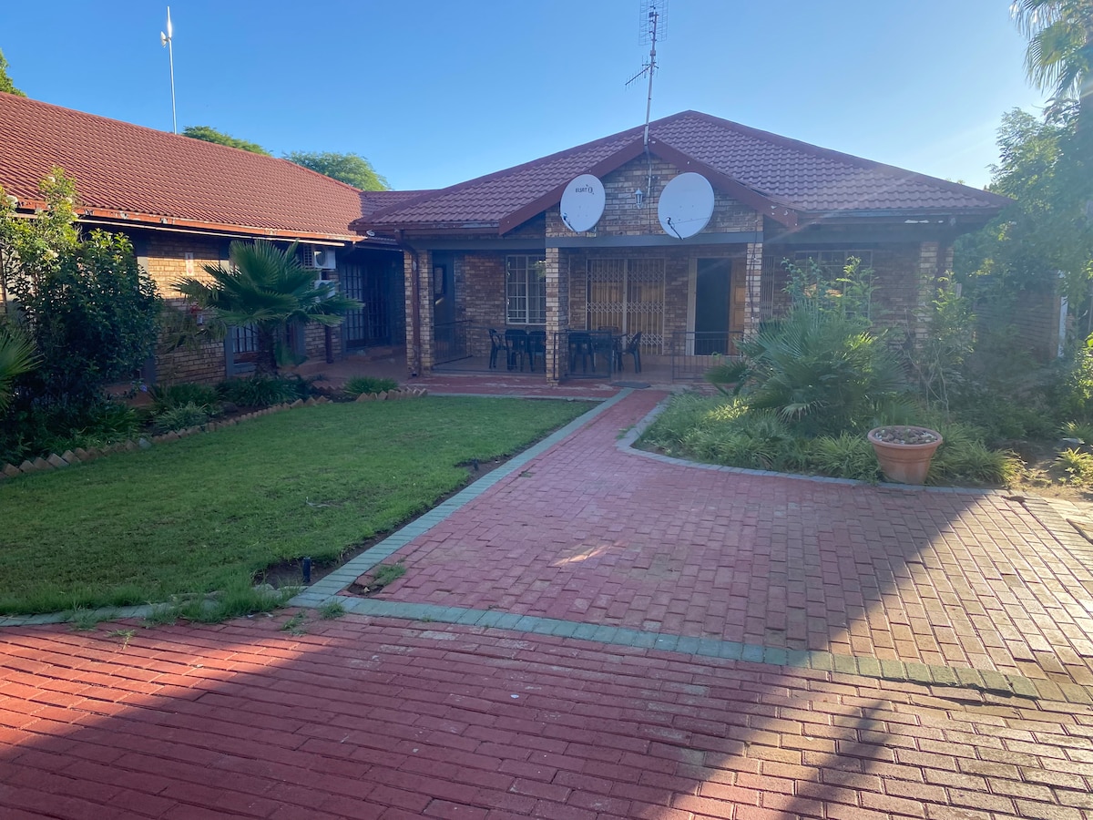 The exterior showcases a charming brick home featuring a well-maintained garden with lush greenery. A paved pathway leads to the entrance, framed by two satellite dishes on the roof. Seating areas are arranged on a covered porch, inviting outdoor relaxation.