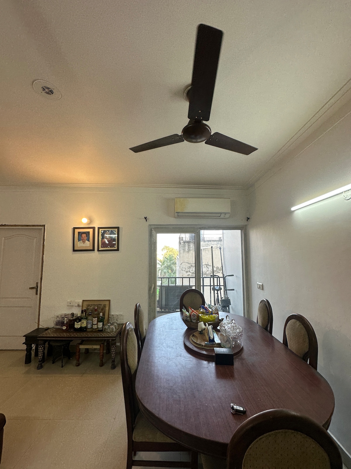 A dining area features a long wooden table surrounded by several chairs. A ceiling fan hangs above, providing ventilation. Wall art is displayed, and natural light enters through a glass door that leads to a small balcony. A tray with glassware sits at the center of the table.