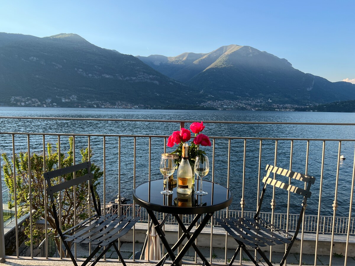 A balcony set for two features a round table adorned with a vase of bright pink flowers. Two glasses and a bottle of wine are displayed, with expansive views of Lake Como and mountains in the background under a clear sky.