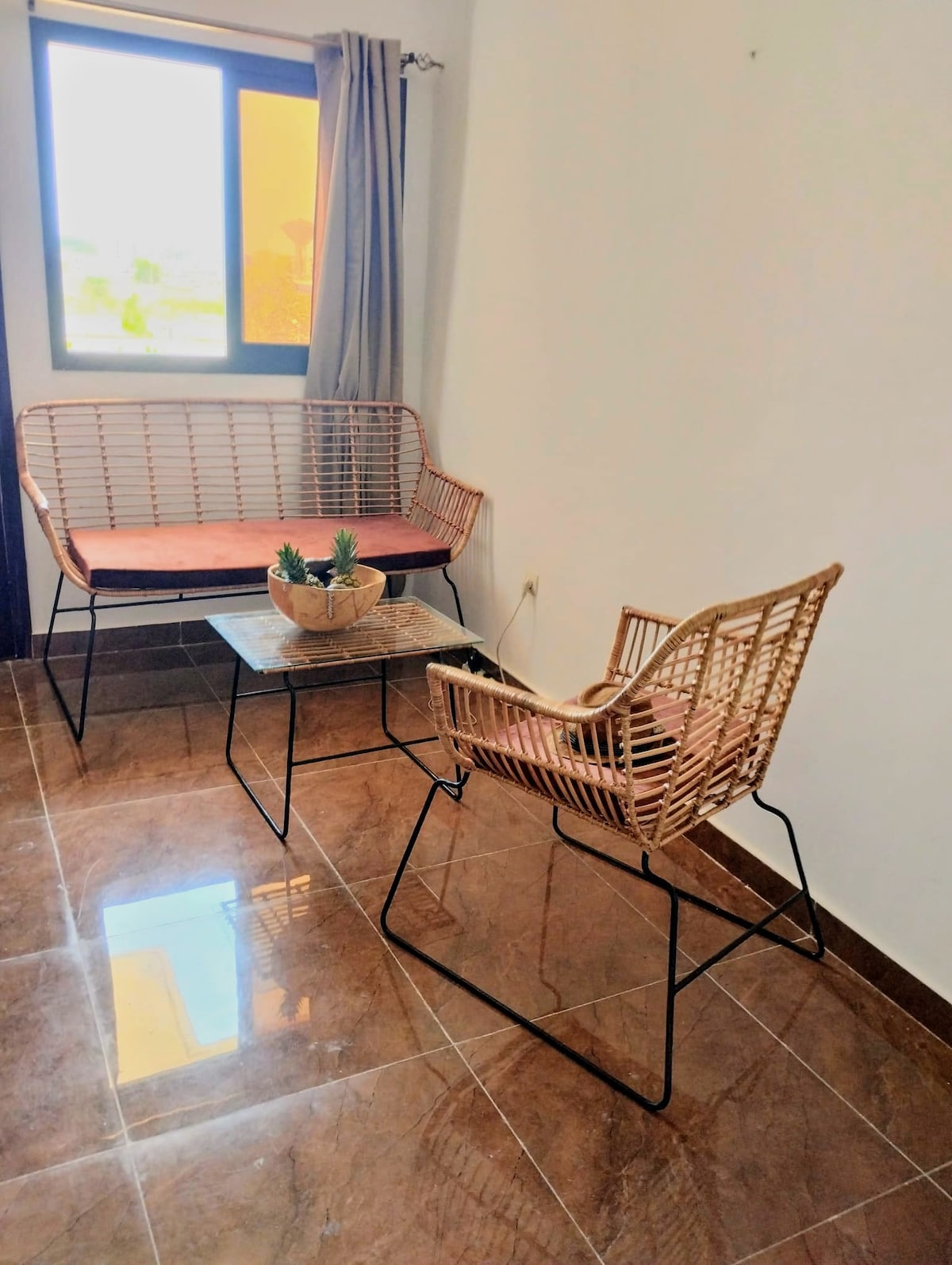 A seating area features a rattan loveseat and an armchair, both positioned around a small glass-topped table. Natural light enters through a window, highlighting the glossy tiled flooring.