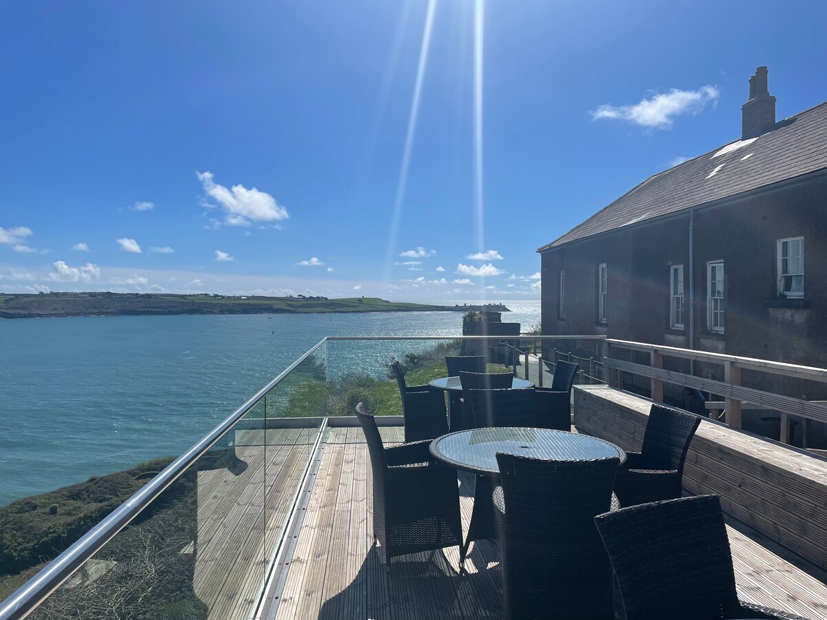 A spacious outdoor terrace features a glass railing overlooking a calm body of water. Several dark wicker chairs surround a circular table, providing a comfortable seating area. Sunlight reflects off the water, and a clear blue sky adds to the serene atmosphere.