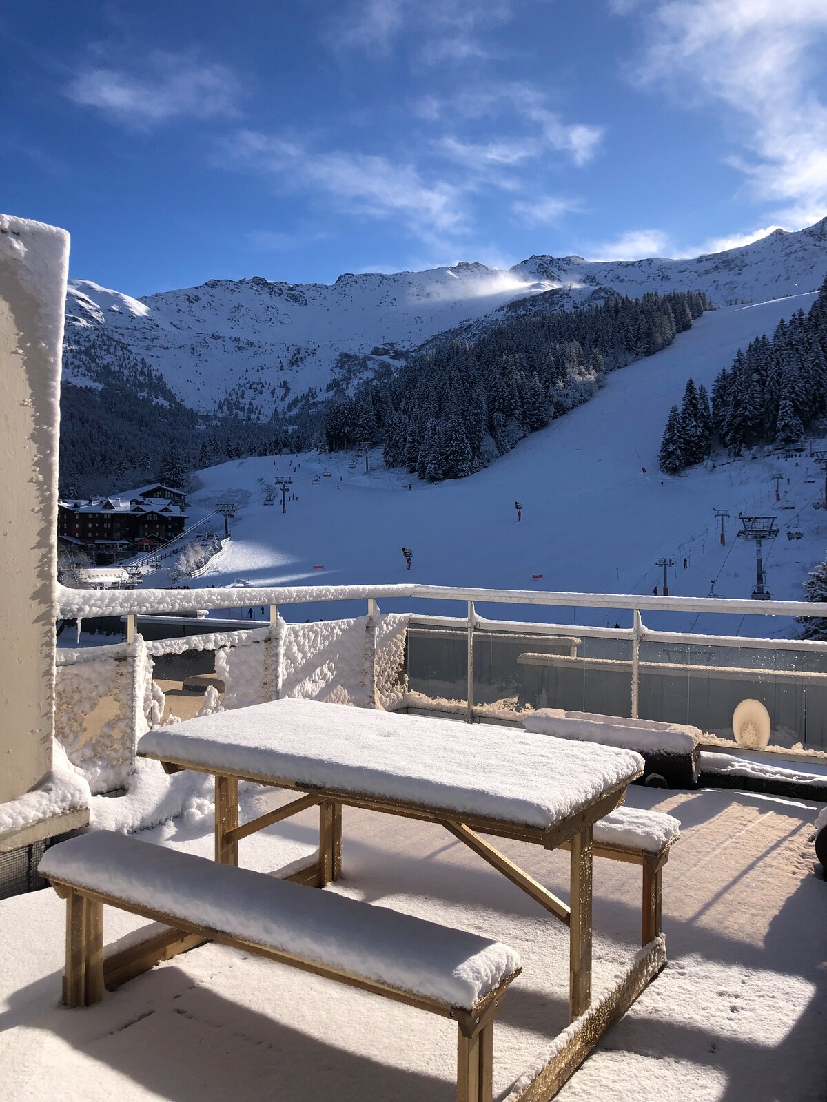 A spacious terrace is shown covered in fresh snow, featuring a wooden picnic table surrounded by snowy views of the mountains. The ski slopes are visible in the distance, with clear blue skies above, enhancing the serene winter landscape.