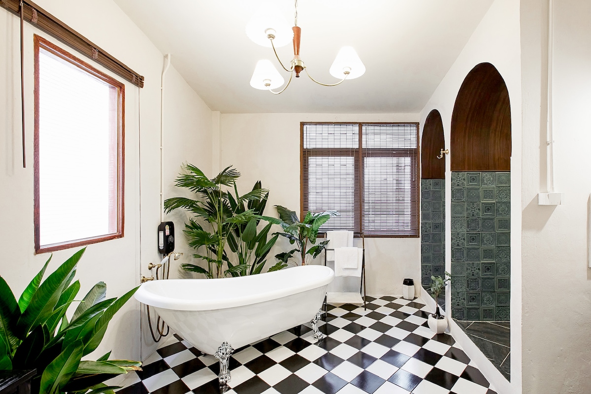 A spacious ensuite bathroom features a freestanding bathtub and a separate rain shower. Tall plants add a touch of greenery, while the floor is adorned with black and white tiles. Natural light filters through the two windows, creating a bright and inviting atmosphere.