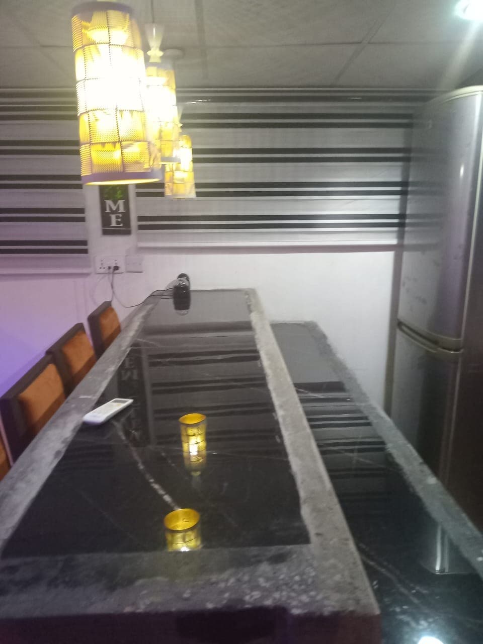 A modern kitchen area features a long black marble countertop with reflective surfaces. Above, pendant lights provide ambient lighting. A sleek refrigerator is positioned nearby, and a set of dark chairs is arranged along the countertop, enhancing the functional design of the space.