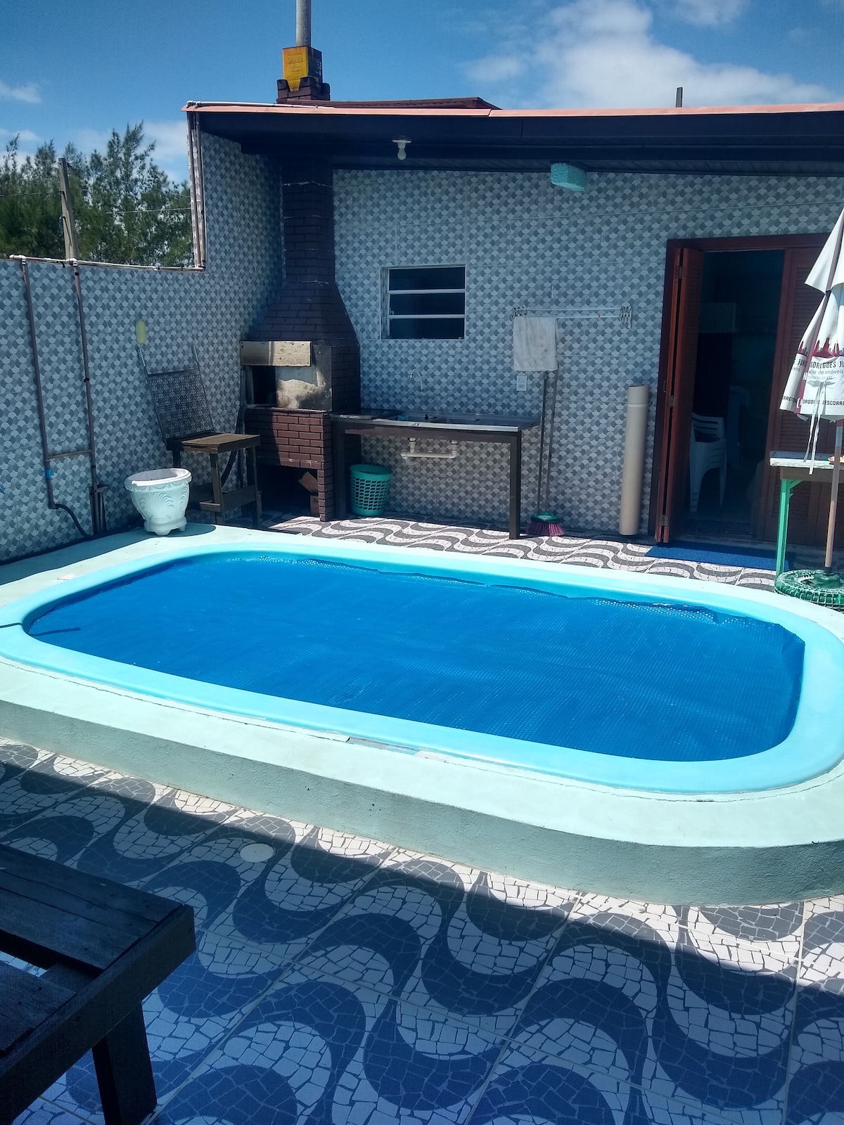 A private outdoor area features a heated pool surrounded by patterned tile flooring. A barbecue grill is visible alongside the pool, and an umbrella provides shade for nearby seating. The wall displays a light tile pattern, contributing to the open atmosphere.