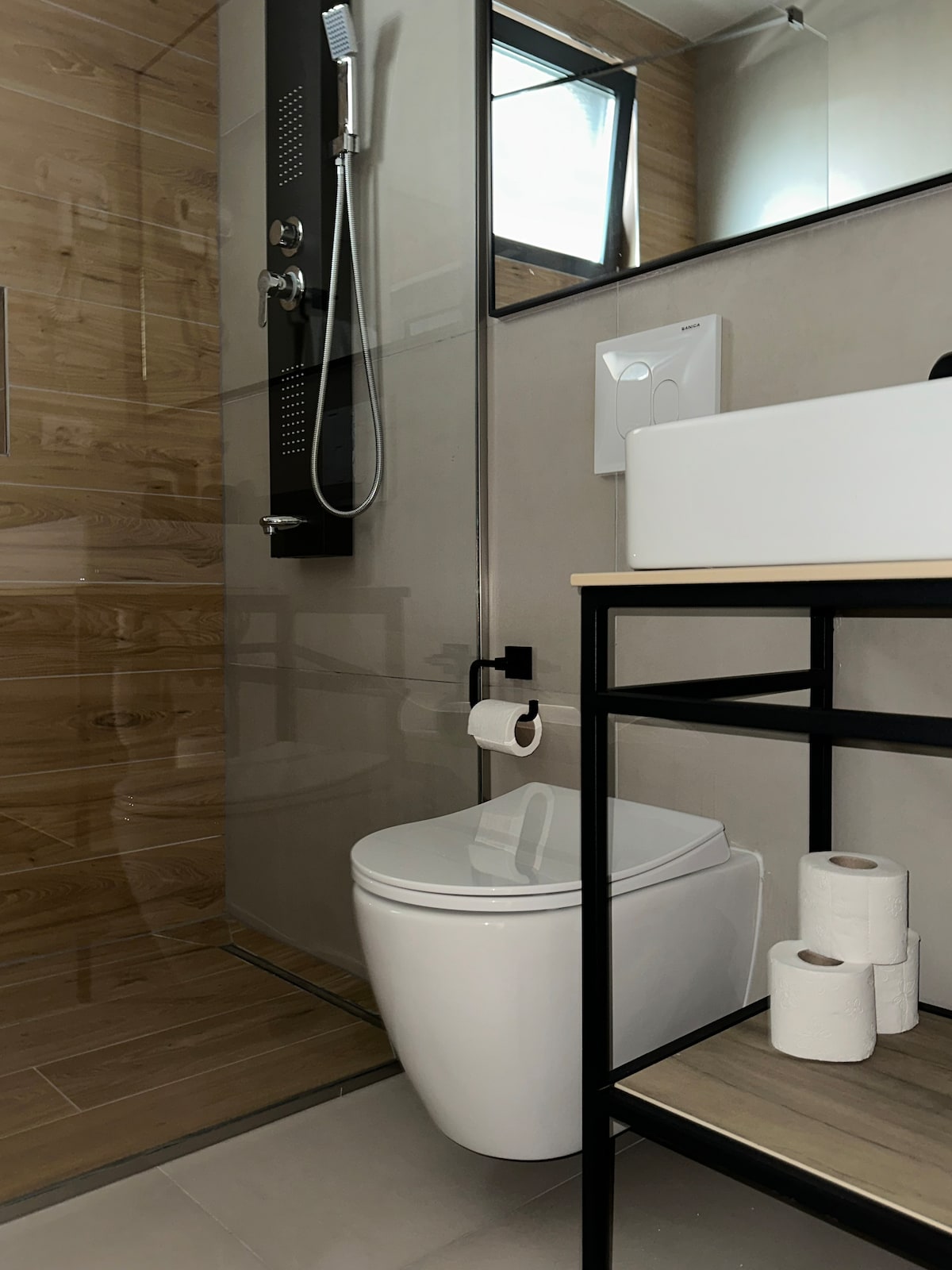 A modern bathroom features sleek, minimalist design elements. A glass-enclosed shower with a handheld showerhead is visible alongside a white sink mounted on a black frame. A toilet is positioned strategically, and toilet paper rolls are neatly stacked on the lower shelf.