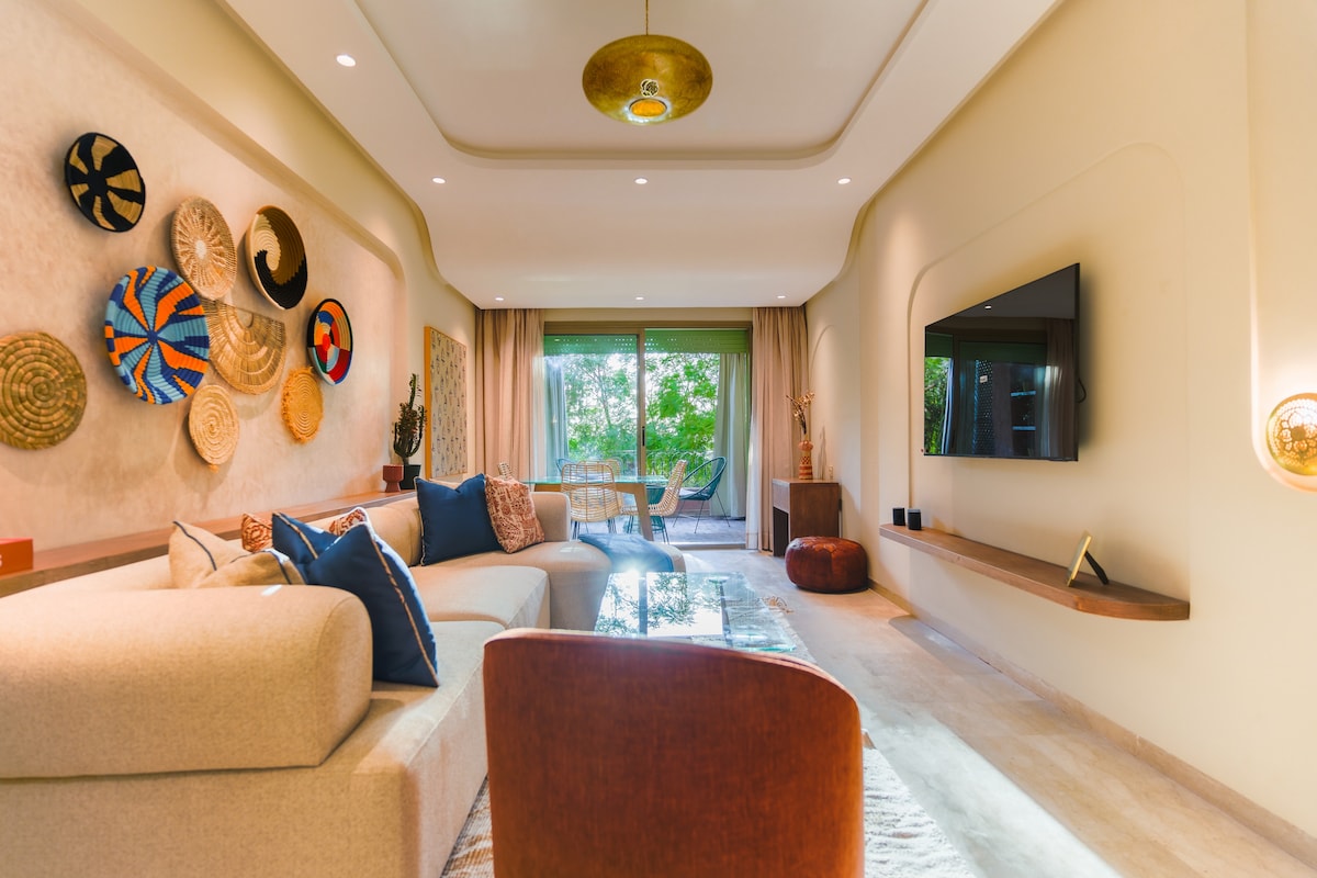 The living area features a light-toned sectional sofa and a sleek coffee table. Textured wall art is displayed, while large glass doors provide access to outdoor views. A flat-screen TV is mounted on a neutral wall, and elegant lighting fixtures add subtle illumination.