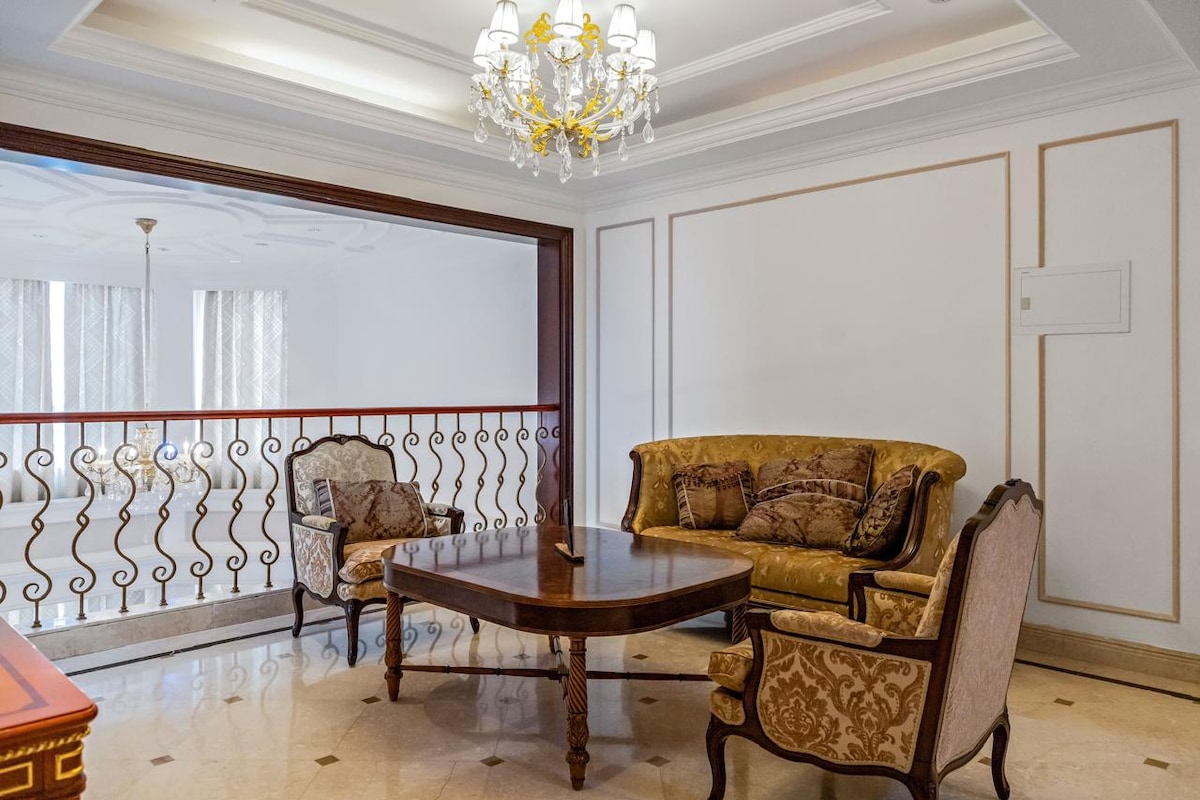 Elegant seating is arranged around a dark wooden coffee table in a well-lit living area. The decor features a classic chandelier overhead and ornate patterns on the armchairs, complemented by a harmonious color palette. A balcony railing can be seen in the background.
