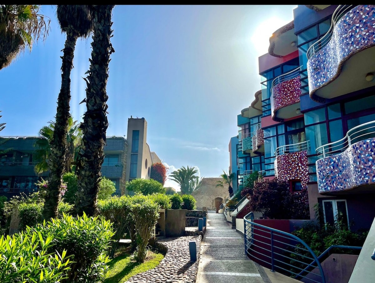 A pathway lined with greenery connects colorful buildings displaying unique architectural features. Sunlight filters through the palm trees, creating an inviting atmosphere. The vibrant colors contrast with the blue sky, enhancing the visual appeal of the serene environment.