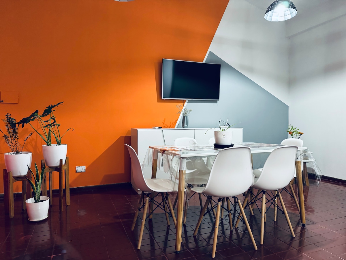 A dining area is highlighted by a stylish table surrounded by six white chairs. An orange accent wall frames the space, complemented by a gray wall. A flat-screen TV is mounted above a white cabinetry unit, while potted plants add a touch of greenery to the setting.