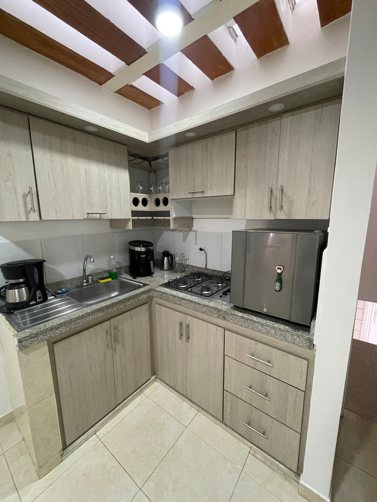 A modern kitchen is outfitted with wood cabinetry and grey granite countertops. Included are essential appliances such as a coffee maker, kettle, and refrigerator. Natural light is provided by overhead fixtures, illuminating the space and highlighting the tiled flooring.
