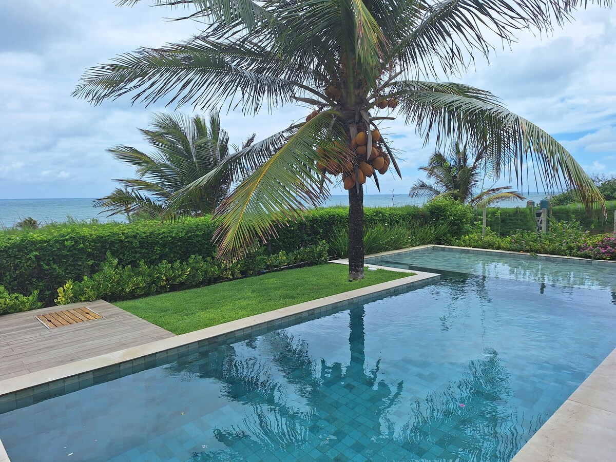 A serene outdoor space features a clear turquoise pool bordered by lush greenery and palm trees. The ocean can be seen in the background, with gentle waves creating a calming backdrop. A wooden deck complements the landscaped area, enhancing the tranquil environment.