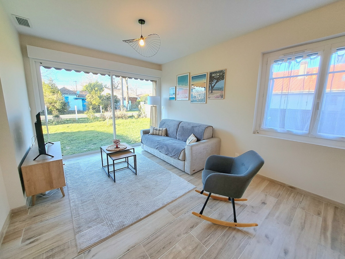 A light-filled living area features a comfortable grey sofa and a rocking chair. A coffee table is positioned centrally on a soft area rug. Large sliding glass doors offer a view of the garden, enhancing the space's openness.