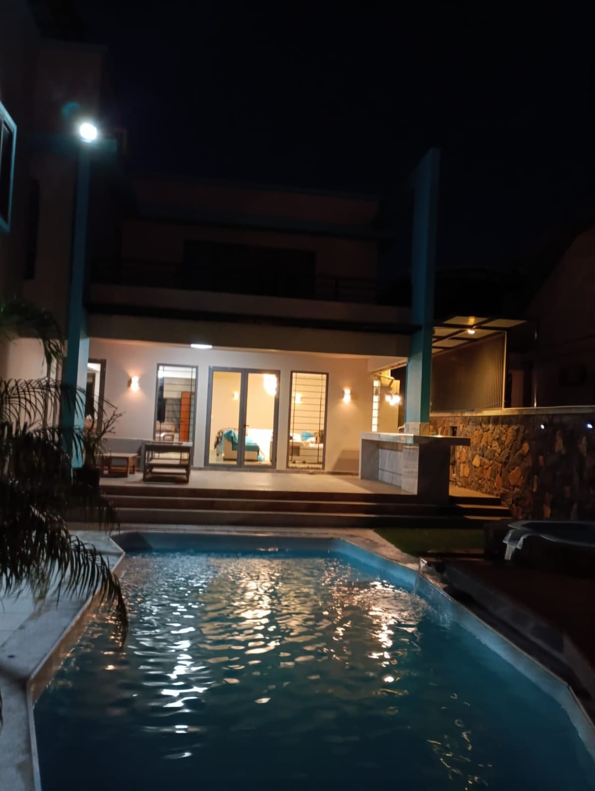 A tranquil outdoor area features a serene swimming pool, illuminated by subtle lighting, reflecting the night sky. Comfortable seating is arranged beside the pool, with a stone wall adding texture and architectural interest to the setting.