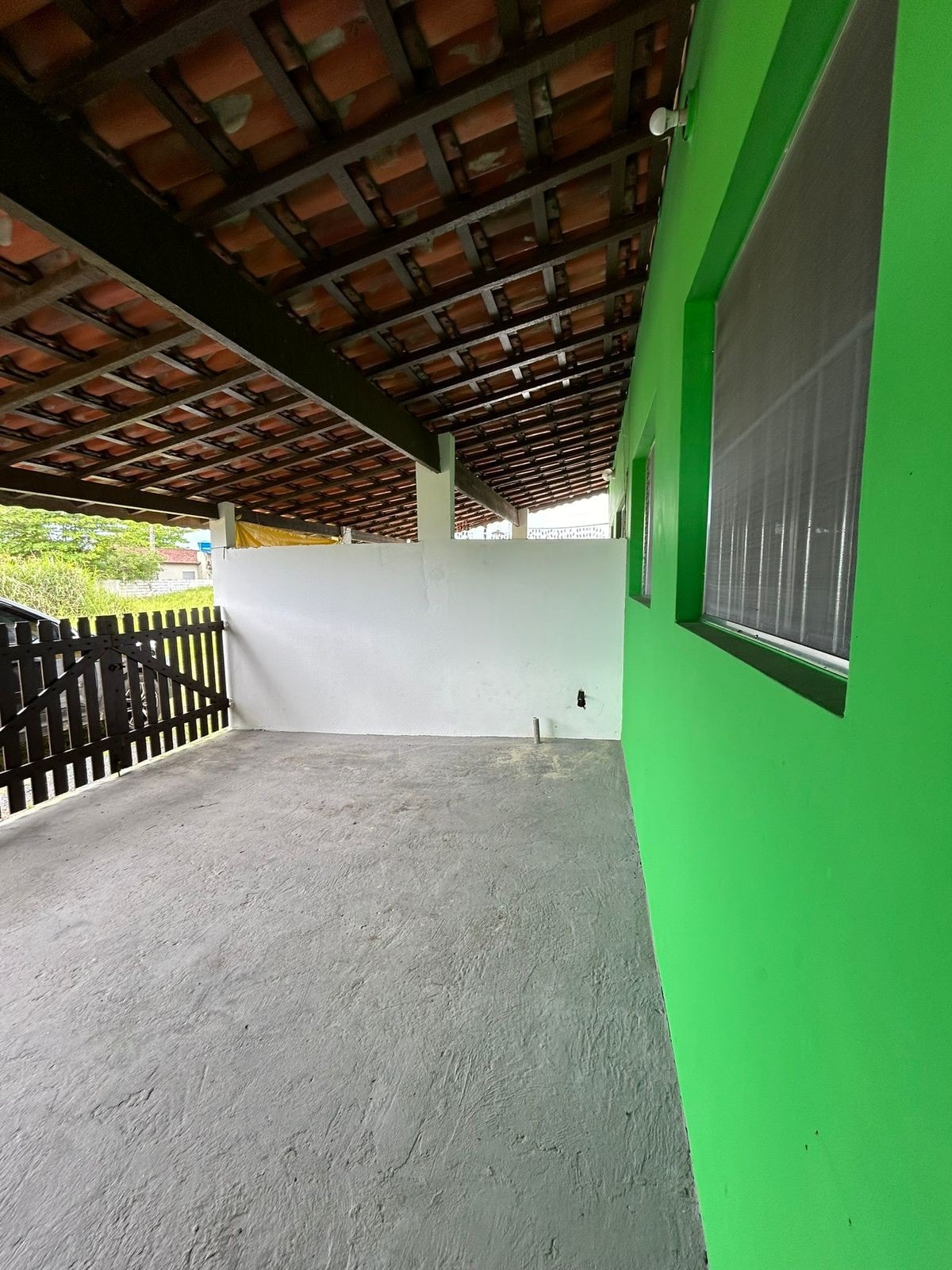 The image displays a covered outdoor area featuring a concrete floor and green walls. A wooden fence is visible, enhancing privacy. The ceiling is supported by dark beams, with terracotta tiles above. Natural light enters through a nearby window, illuminating the space.