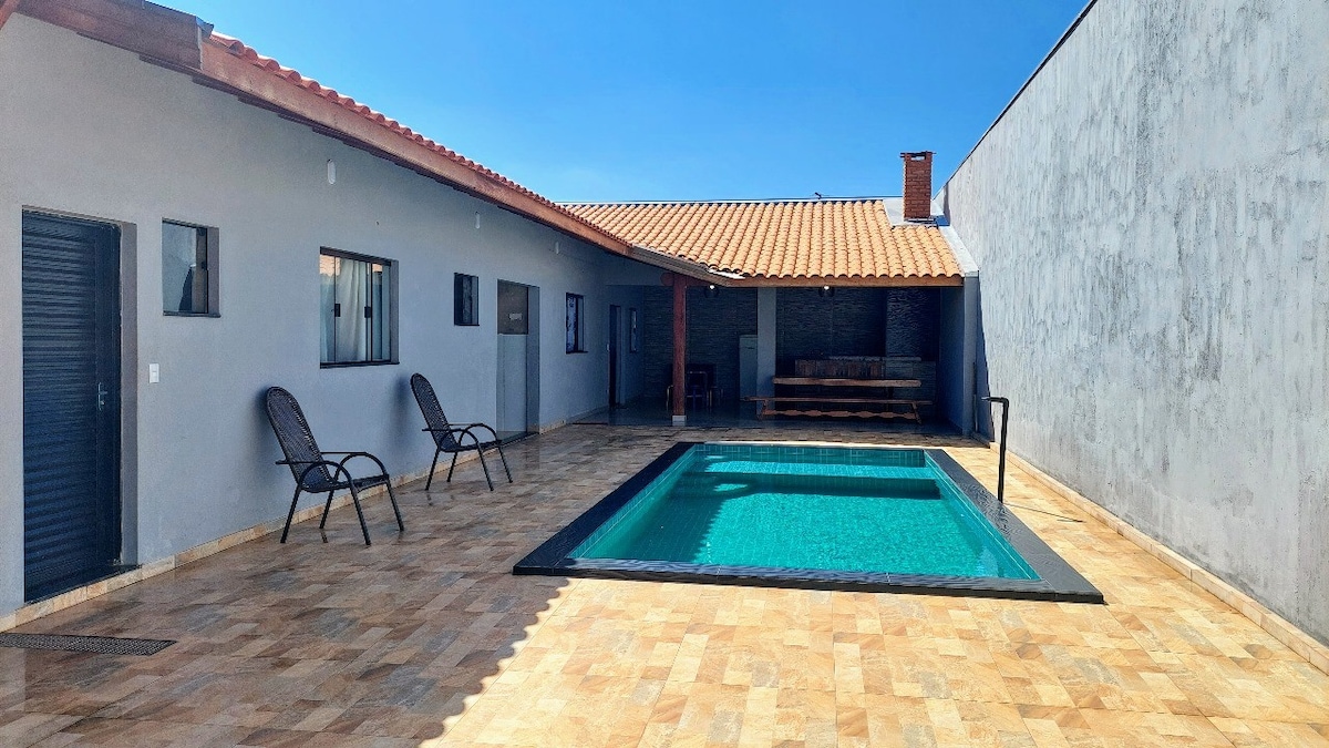 A spacious outdoor area features a rectangular swimming pool surrounded by a tiled patio. Loungers are positioned nearby, and a shaded seating area is visible against the back wall with wooden benches. The structure has multiple windows, allowing natural light to enter.
