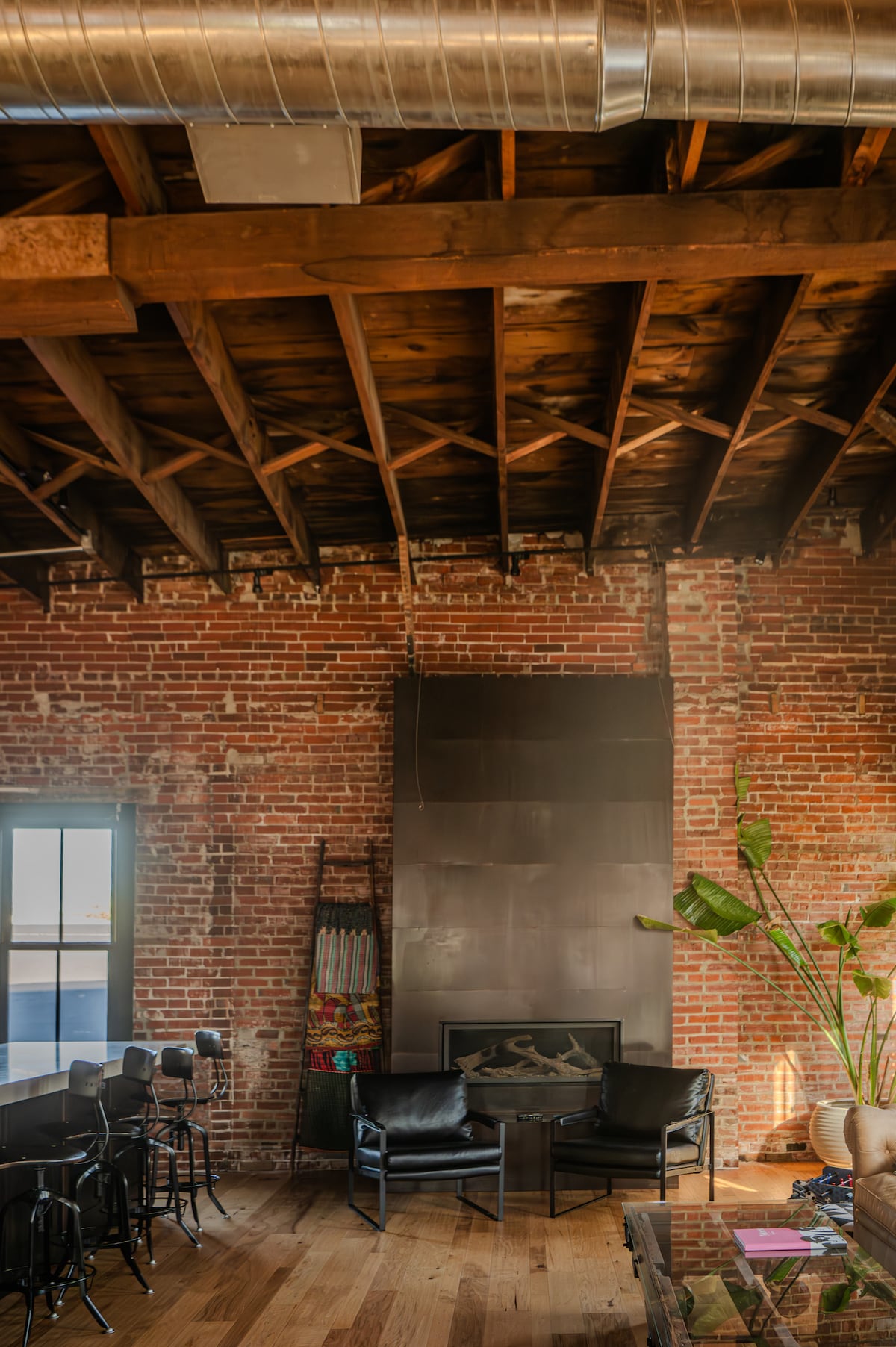An inviting lounge area is highlighted by exposed brick walls and a high wooden ceiling. Two black chairs are positioned around a sleek fireplace, with a potted plant adding a touch of greenery. Large windows allow natural light to enhance the space.