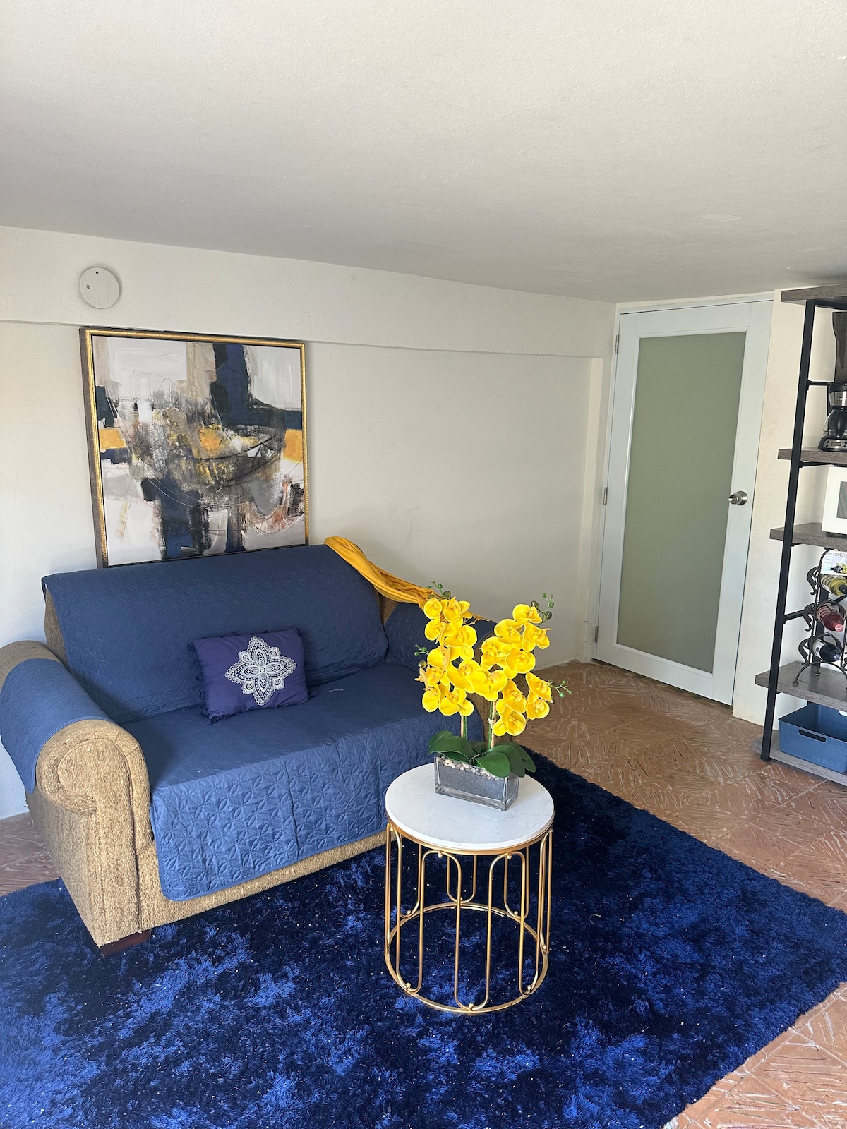 A cozy seating area features a blue sofa adorned with a decorative pillow. A round coffee table with a gold base holds a vibrant arrangement of yellow flowers. A colorful wall art piece is displayed above the sofa, complemented by a partially open door leading to another room.