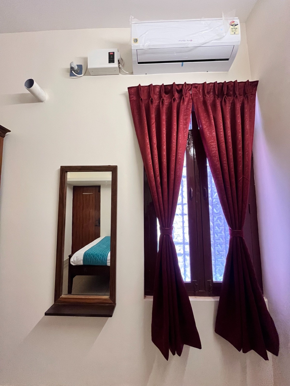 A wall-mounted air conditioner is positioned above a wooden mirror, reflecting a window adorned with rich maroon curtains. The light-colored walls contribute to a bright and airy feel in the room, inviting natural light to enhance the space.