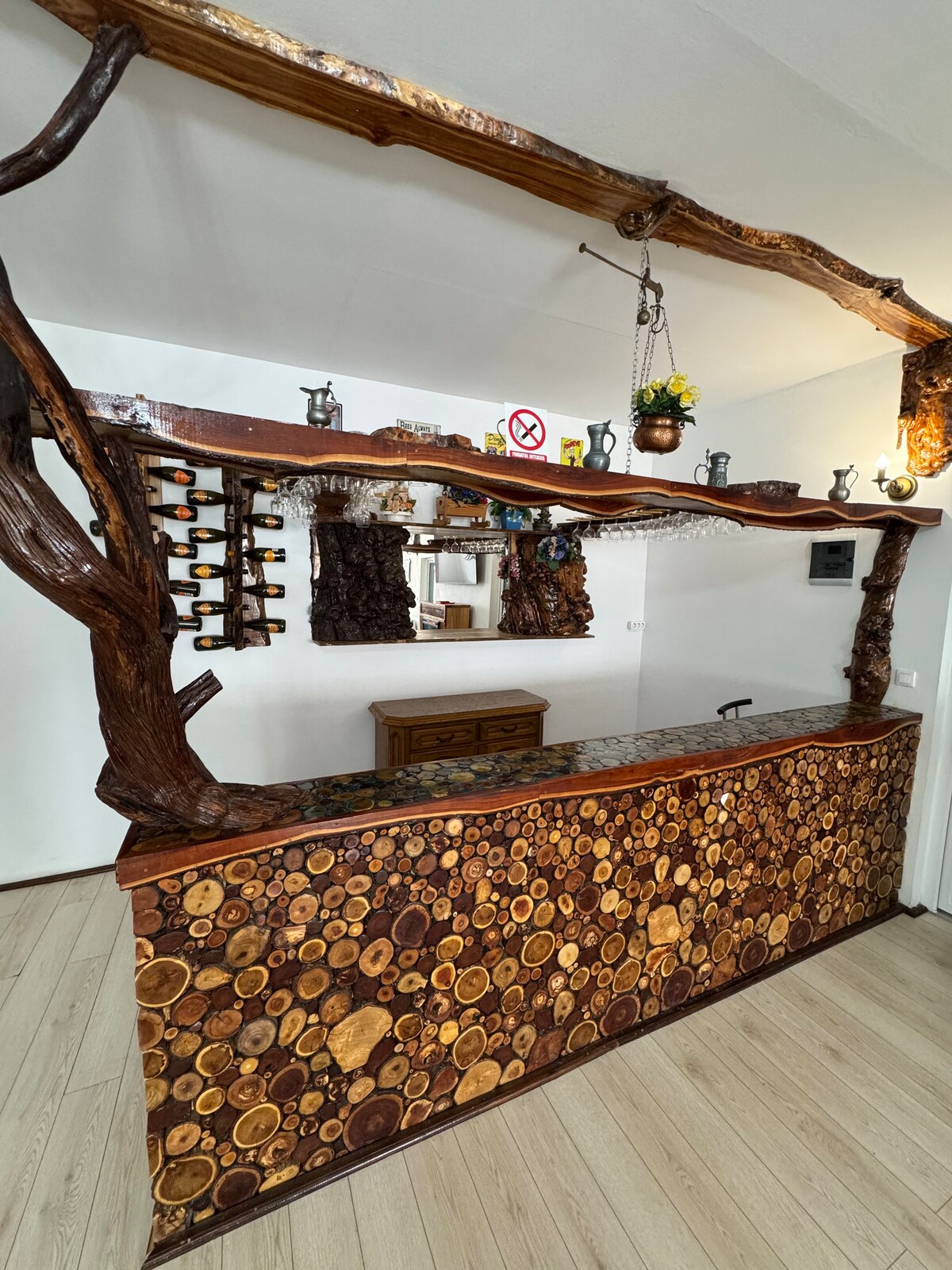A unique reception area features a wooden counter adorned with circular logs, showcasing natural textures. Above the counter, decorative elements include hanging plants and metal vessels. A mirror reflects the warm tones of the wood, contributing to an inviting atmosphere.