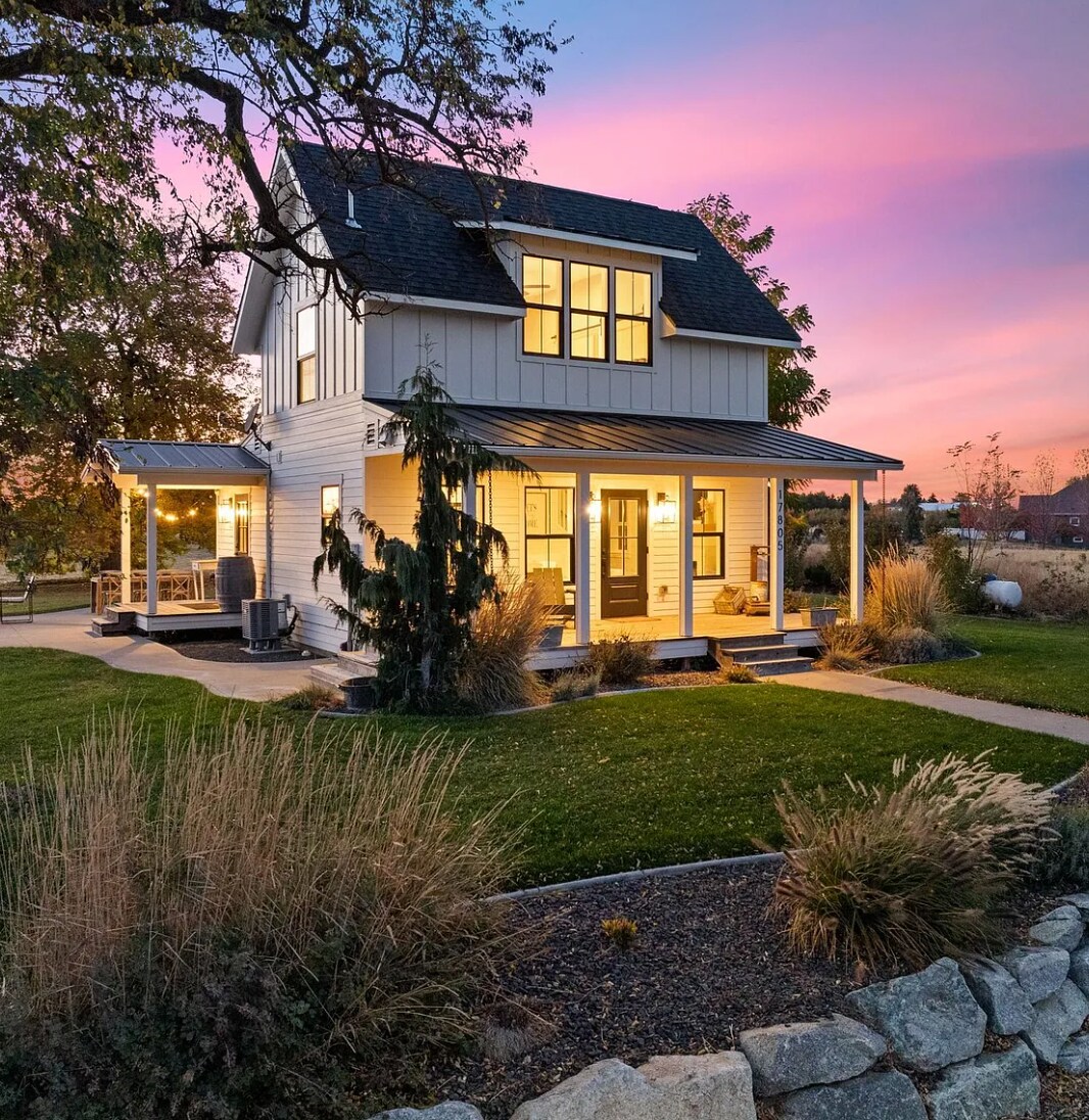 A charming two-story cottage is set against a vibrant sunset sky. The exterior features a welcoming porch with seating and large windows that allow warm light to spill out. Lush grass and landscaped areas surround the home, creating a serene outdoor environment.