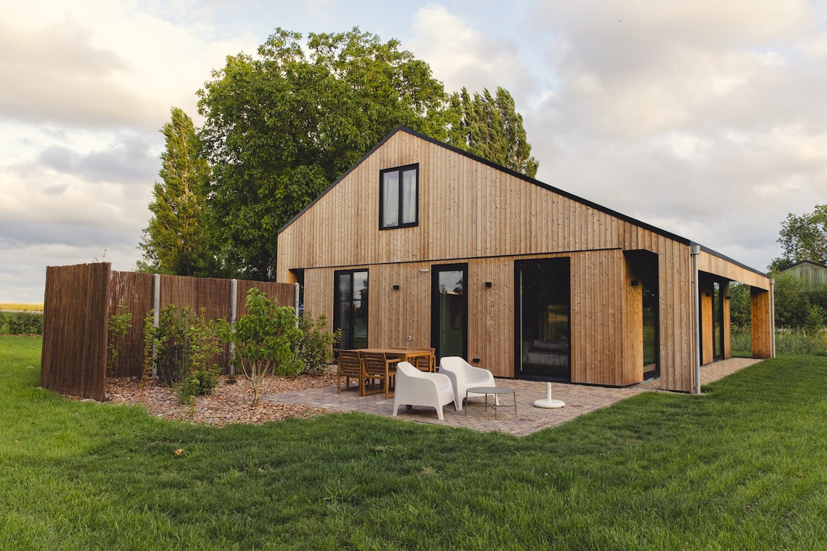 The exterior of a modern wooden structure is displayed, surrounded by green grass. A terrace with outdoor seating is visible in front, and large windows allow natural light to fill the interior. Lush trees are positioned in the background, enhancing the natural setting.