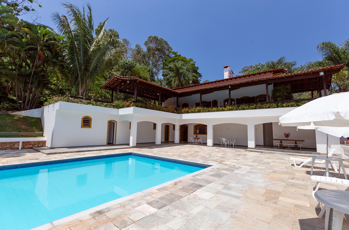 The outdoor area features a swimming pool surrounded by stone flooring. The two-story house overlooks the pool, with large windows and a terraced roof area adorned with greenery. Sun loungers and a shaded umbrella provide relaxation options in the garden setting.