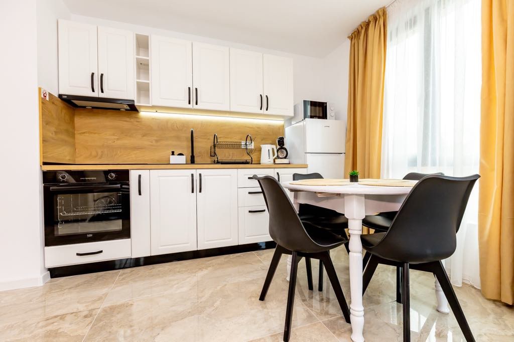 A modern kitchen area features sleek white cabinets and wood accents. A dining table is set for four, surrounded by black chairs. Natural light pours in through a large window, and appliances, including a stove and refrigerator, provide essential amenities.
