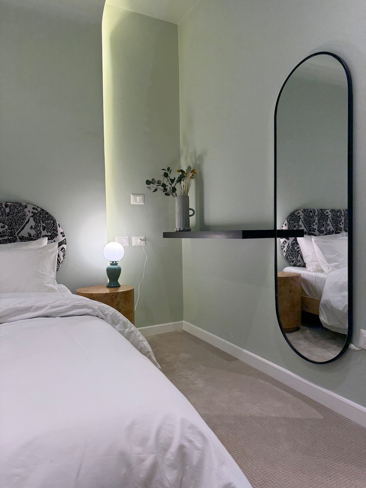 A serene sleeping area features a spacious bed with neutral bedding. A minimalist black mirror reflects the room's calming green walls. A small wooden nightstand holds a round lamp, complemented by a vase with dried flowers on the shelf above.