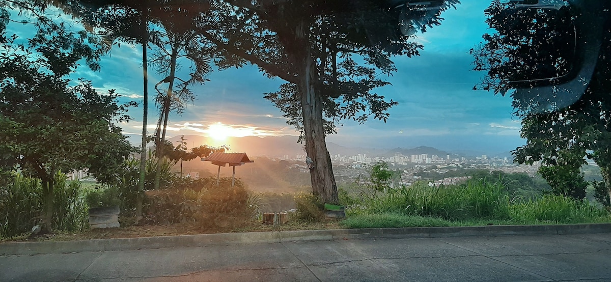 A scenic view captures a sunset behind distant mountains, with soft, colorful sky tones reflected in the clouds. Lush greenery surrounds the area, while a small structure is visible in the foreground, providing a place to enjoy the landscape.