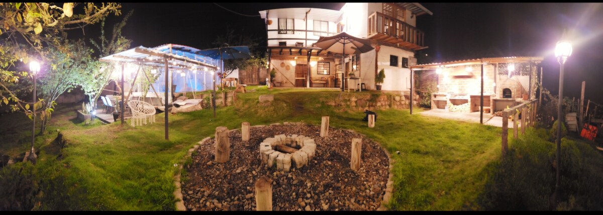 The outdoor area features a circular fire pit surrounded by stone seating, with lush grass and scattered trees. Twinkling lights adorn the nearby structures, including a covered porch and a seating area, creating a serene atmosphere for gatherings under the evening sky.