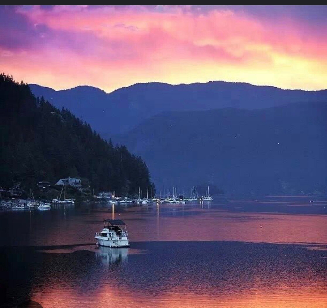 A serene landscape features a calm lake surrounded by mountains at sunset, with soft colors reflecting on the water. A boat is positioned in the foreground, while distant boats are visible along the shoreline amidst lush greenery.