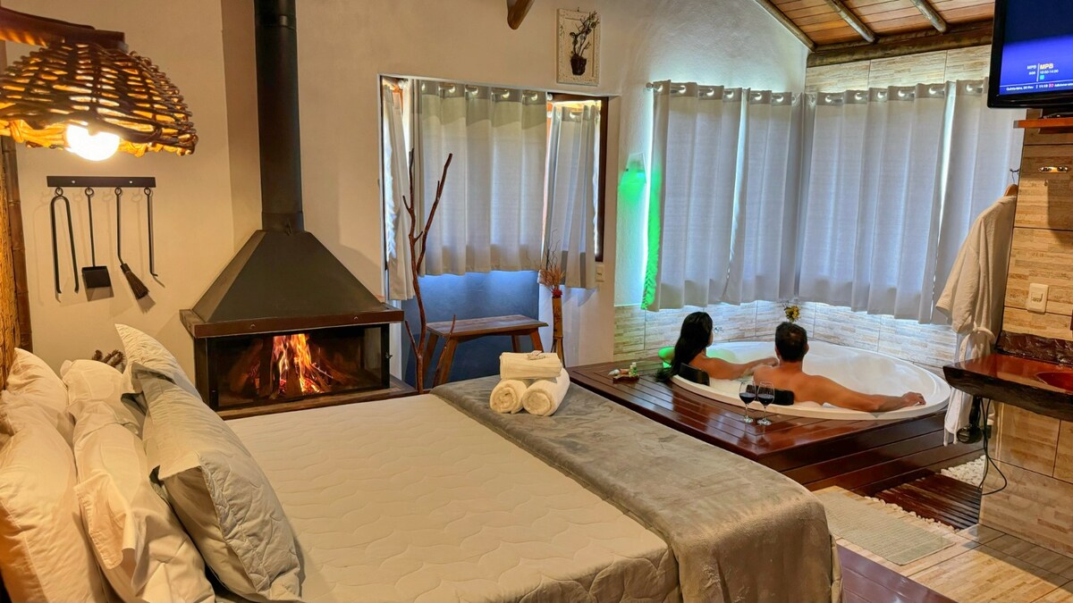 A cozy room showcases a spacious bed with light grey bedding, situated near a modern fireplace. Soft, sheer curtains frame a large window, while a relaxing bathtub is positioned close by. A flat-screen TV is mounted on the wall, enhancing the room's comfort and functionality.