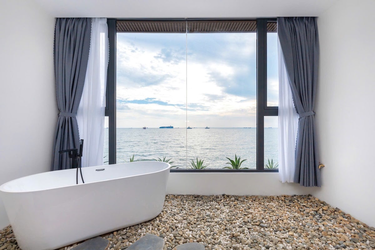 A spacious bathroom features a freestanding tub placed in front of large windows, offering an unobstructed view of the sea. Soft gray curtains frame the windows, while a pebble-stone floor complements the modern design.