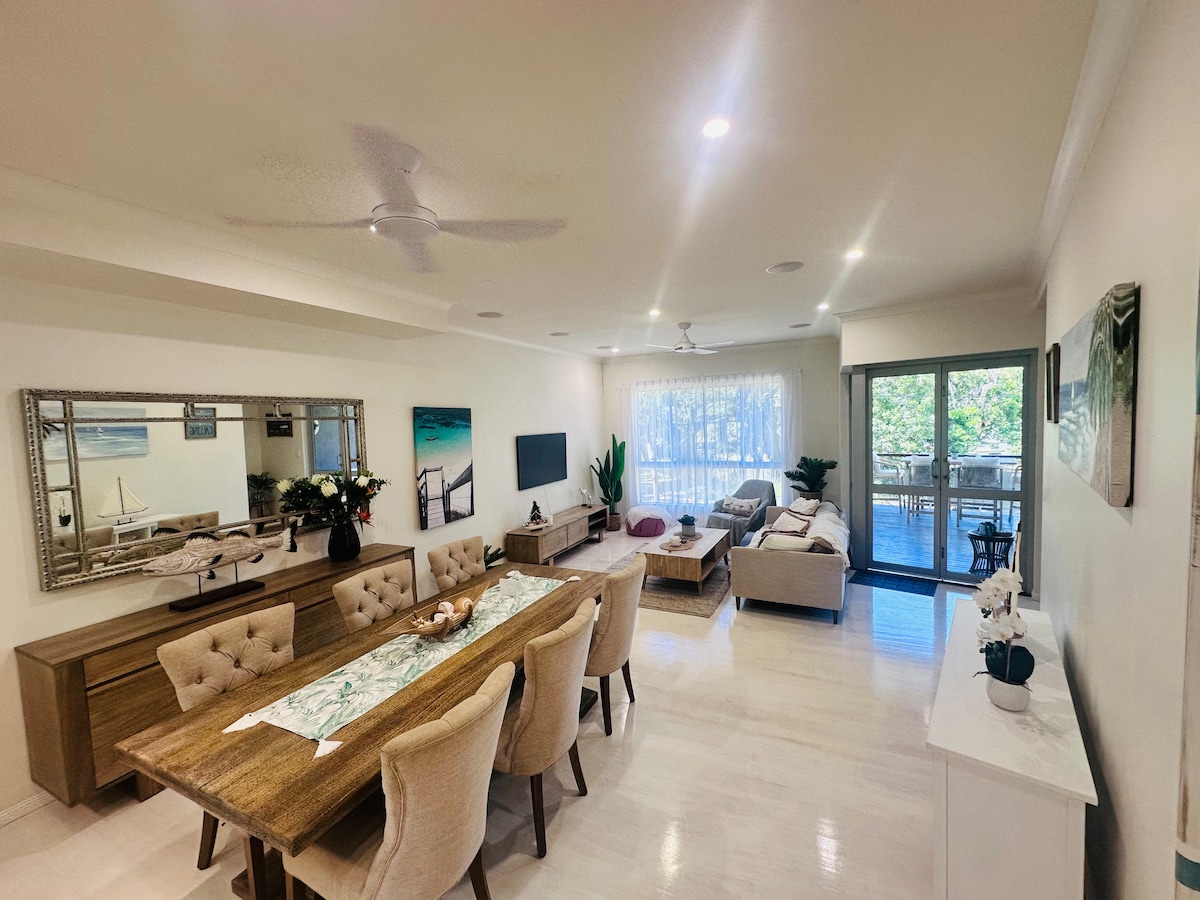 A spacious dining area is featured with a wooden table surrounded by upholstered chairs. Natural light fills the open living space, which includes a comfortable seating area and a television. Sliding glass doors lead to a balcony, enhancing the airy feel.