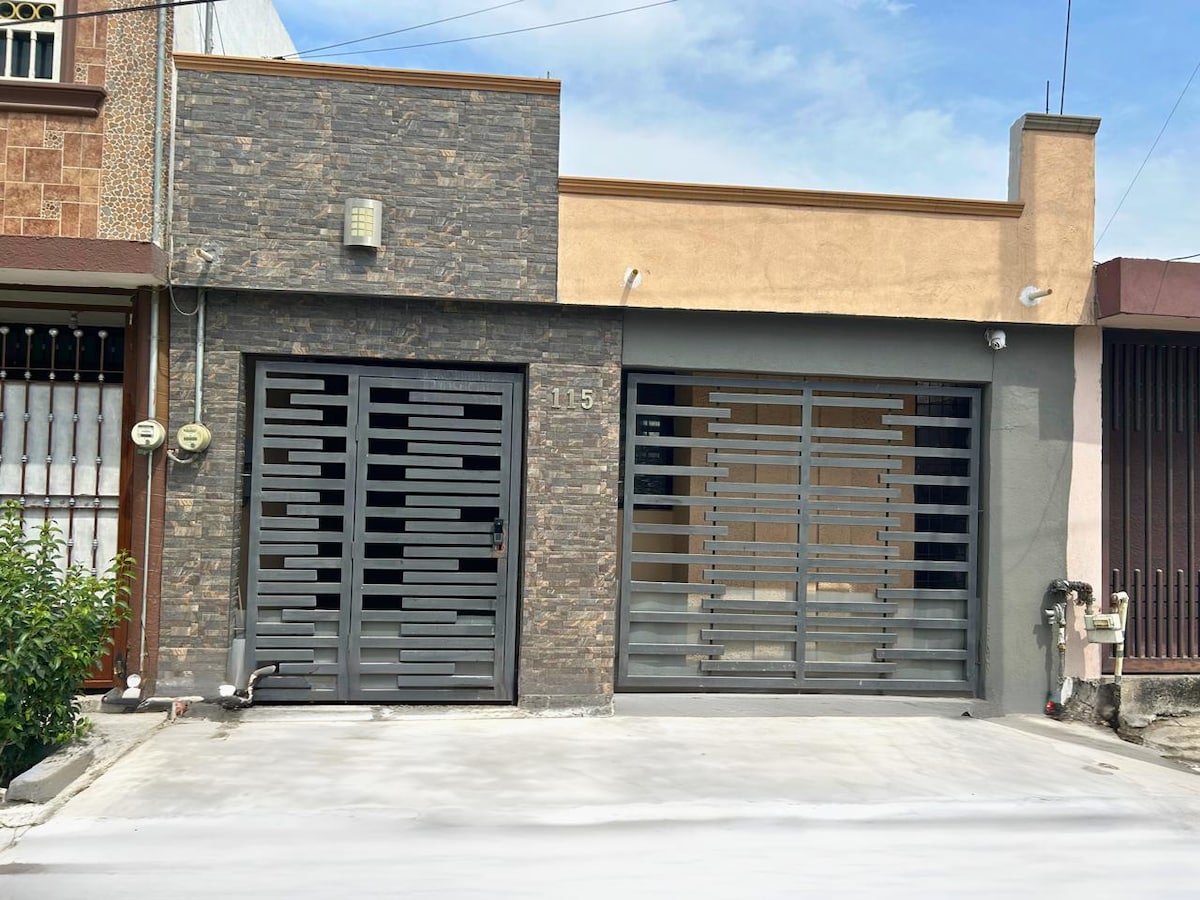 The exterior of the property is depicted, showcasing a modern facade with a combination of stone and smooth surfaces. A contemporary metal gate is visible, allowing entry. The driveway is spacious and leads to the main entrance, framed by neutral-colored walls.