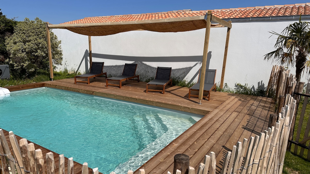 A wooden deck surrounds a heated pool, featuring three sun loungers placed beneath a shaded canopy. The serene water reflects the clear sky above, while a wooden fence adds privacy to the outdoor space, complemented by some greenery in the background.